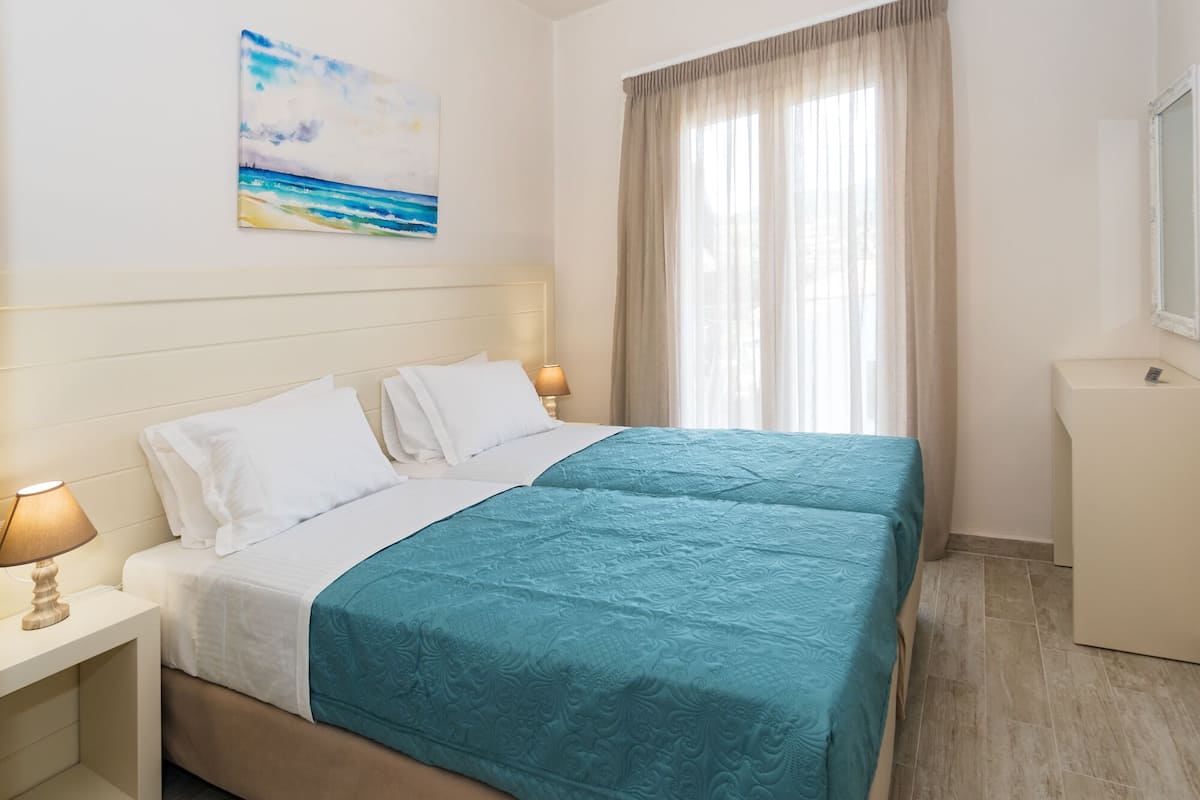 Una habitación de hotel con una cama, dos mesitas de noche, una lámpara, un cuadro, un ventanal con cortinas y un pequeño armario blanco.