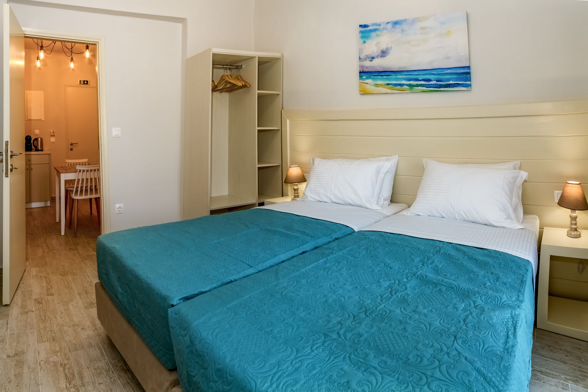 A hotel room with a bed, two bedside lamps, a painting of a beach, and a wardrobe with hangers.
