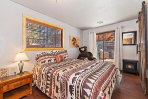 A bedroom with a large bed, a wooden nightstand, a lamp, a teddy bear, a fireplace, and a window with blinds. - Close to Bear Mountain & Golf Course! Hot Tub! Fenced Yard! Foosball! (Big Bear Lake)
