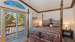 A four-poster bed with a patterned bedspread, a bench at the foot of the bed, a nightstand with a lamp, and a view of the outdoors through a sliding glass door.