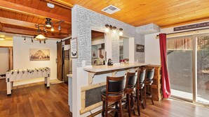 A cozy kitchen with a wooden bar and stools, a foosball table, and a large window with a red curtain.