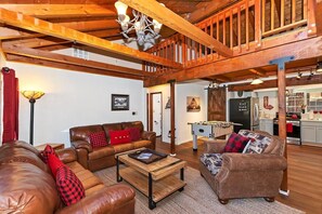 A spacious living area with a high ceiling, wooden beams, a fireplace, and a kitchen in the background.