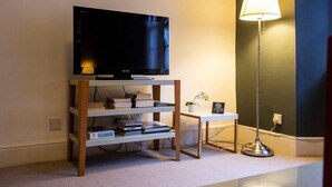 Ein Zimmer mit Fernseher auf einem Holzboden, einem kleinen Tisch mit Lampe sowie einem Regal mit Büchern und einer Pflanze.