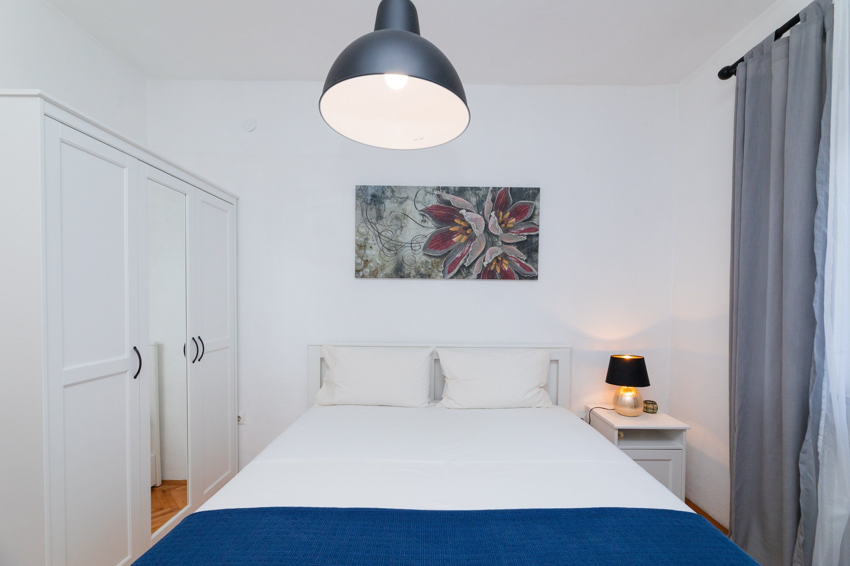 Camera da letto con un letto, un armadio bianco, un quadro, una lampada da comodino e una finestra con tende.