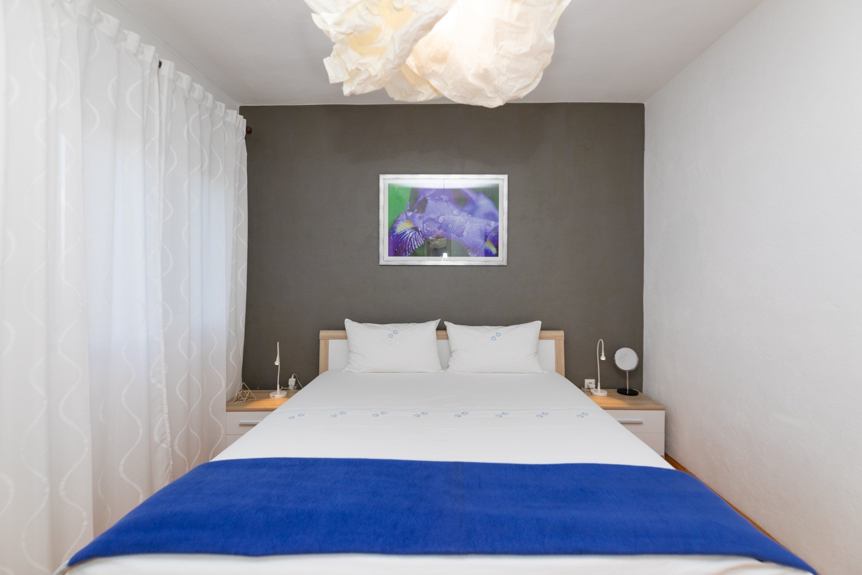 A neatly arranged bedroom with a bed, white curtains, a wall-mounted picture, bedside tables with lamps, and a blue blanket.