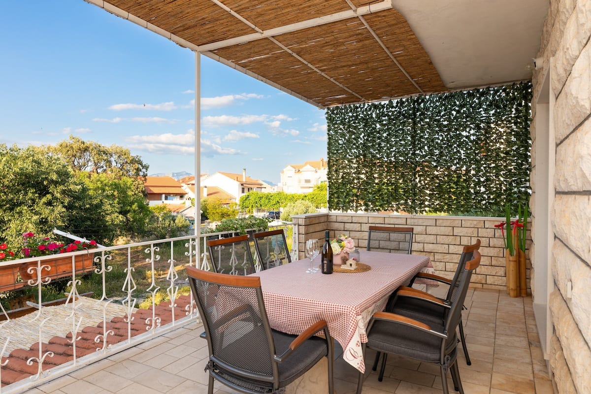A balcony with a table set for two, surrounded by chairs, a pergola overhead, and a view of the neighborhood.