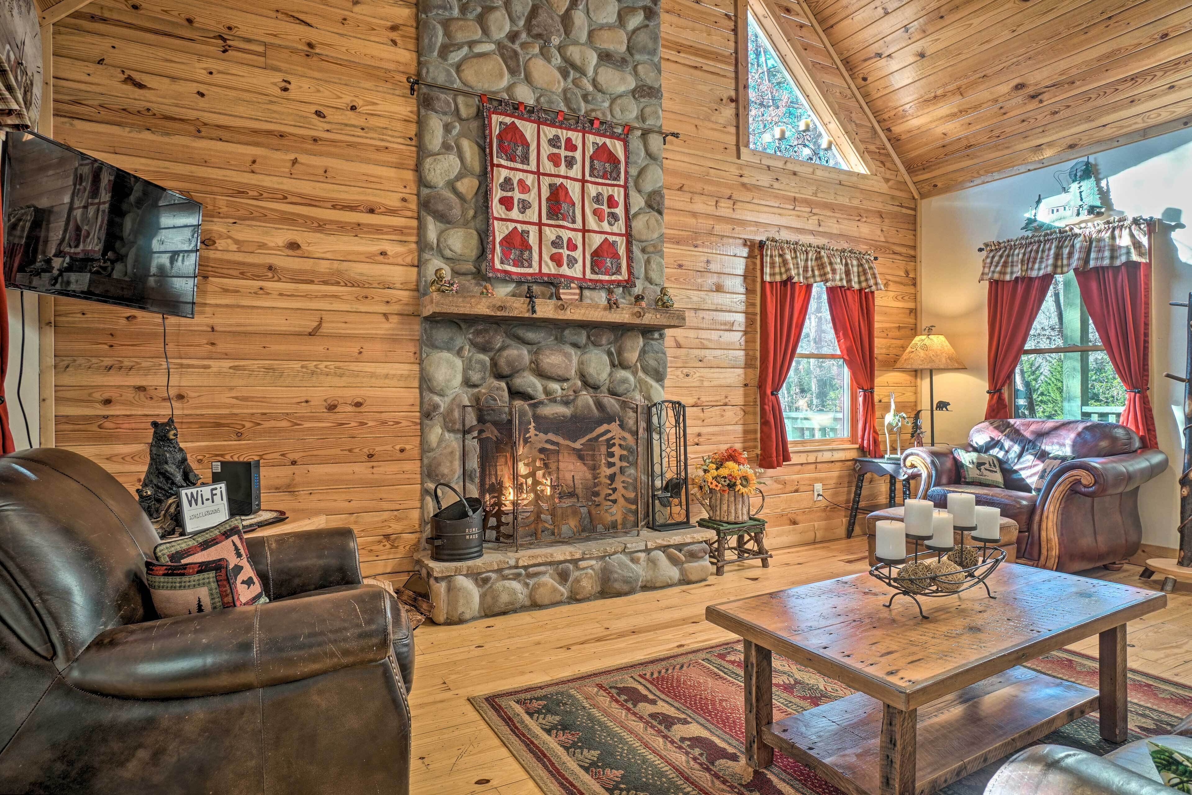 L’intérieur d’un chalet confortable, avec des murs en rondins de bois, une cheminée en pierre, des meubles en cuir et une grande fenêtre agrémentée de rideaux.