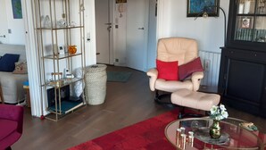 A living room with a red sofa, a beige armchair, a glass coffee table, and a red rug. There is a bookshelf with various items and a door in the background.