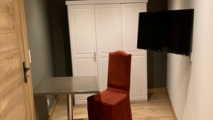 Una habitación pequeña con piso de madera, una silla roja, una mesa con lámpara, un armario blanco y un televisor fijado en la pared.