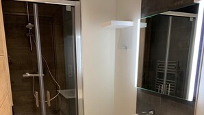 Un baño moderno con cabina de ducha de vidrio, tocador blanco con espejo y suelo de baldosas oscuras.