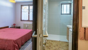Un dormitorio con cama, mesita de noche, ventana con cortinas y baño con ducha e inodoro.