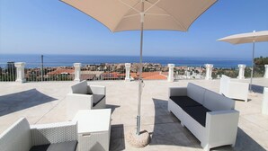 Une terrasse dotée de meubles en osier blanc et de parasols beige, offrant une vue sur le paysage urbain de la côte.