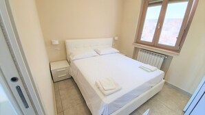 Une chambre avec un lit simple, une table de chevet, une fenêtre avec des stores et un radiateur.