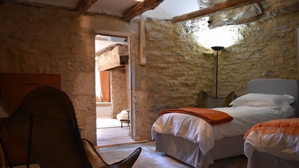 Quarto rústico com paredes de pedra aparente, teto de madeira, cama com roupa de cama branca e manta laranja, e uma poltrona de couro.
