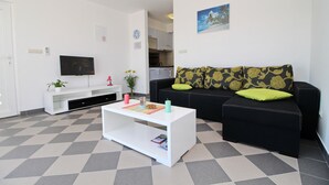 Una sala de estar con una mesa de centro blanca, sofá negro con cojines estampados, un televisor de pantalla plana y una zona de cocina al fondo.