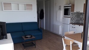 Ett kompakt vardagsrum med en blå soffa, ett litet träbord, en liten köksdel med mikrovågsugn och ugn samt ett vitt skåp.