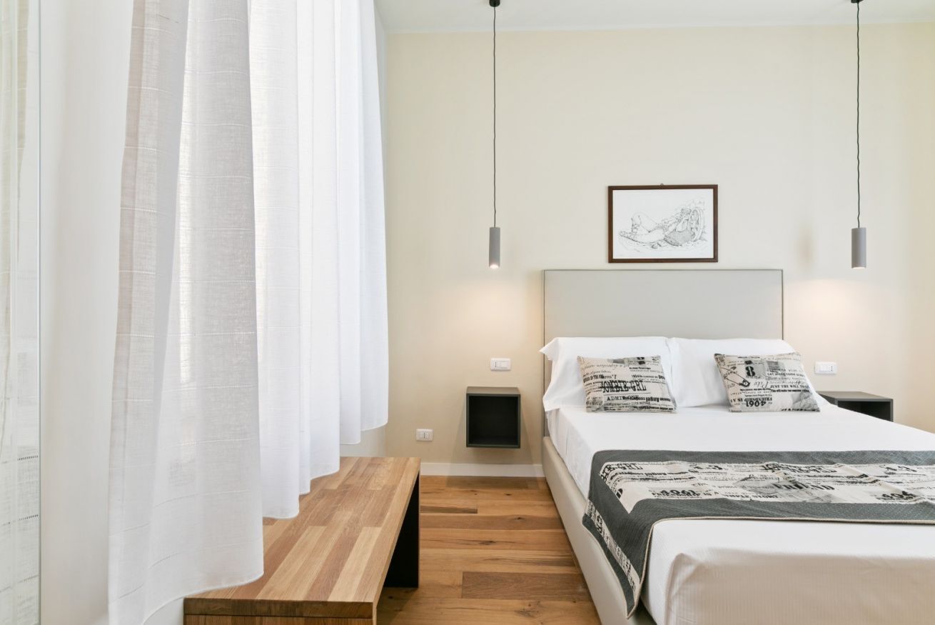 Quarto com cama, mesas de cabeceira, um banco, um quadro na parede e uma luminária de teto.