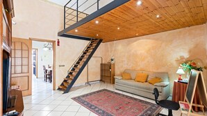A spacious living area with a staircase, a patterned rug, a sofa with cushions, a desk with a chair, and a small table with a lamp.