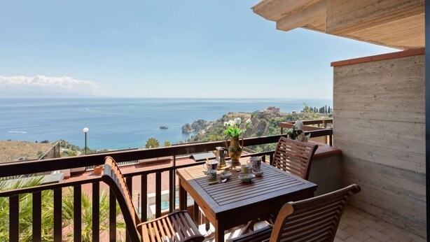 A balcony with a wooden table and chairs, overlooking a coastal view with a clear sky. - Taormina Panoramic Seaview Apartment (Taormina)