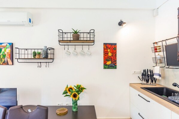 A modern kitchen with a built-in stove, a sink, and a dining area with a table and chairs. There are wall-mounted shelves with plants and a painting.