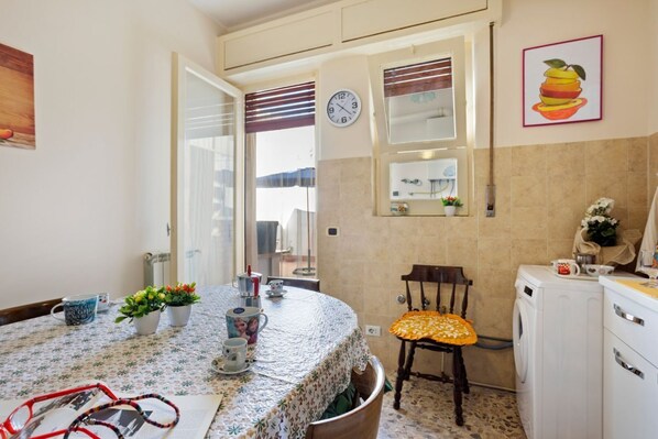 A kitchen with a round table, chairs, a clock, a washing machine, and a window with blinds.