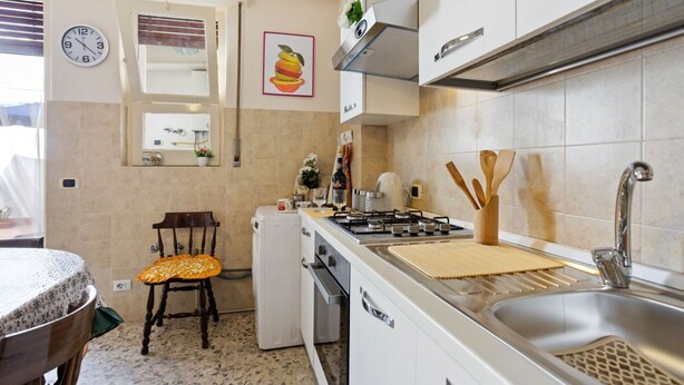 Een keuken met een fornuis, spoelbak en kastjes. Er staat een stoel met kussen, een koelkast, en een klok aan de muur.