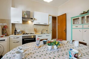 A kitchen with white cabinets, a stove, and a sink. There is a dining table with a floral tablecloth and several mugs.