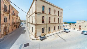 Une rue bordée d’un immeuble dont l’entrée se trouve à l’angle, avec une moto garée devant, et une vue dégagée sur la mer en arrière-plan.