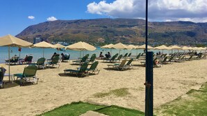 Una playa con filas de tumbonas y sombrillas, cielo despejado y un paisaje montañoso al fondo.