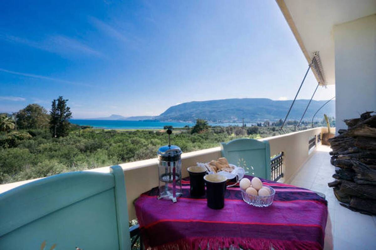 Un balcon avec une table dressée pour le petit-déjeuner, offrant une vue panoramique sur un paysage magnifique, avec des montagnes et une étendue d’eau.
