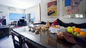 Una sala de estar con una mesa de centro de vidrio, un sofá y un televisor. Hay pinturas en la pared y una ventana con vistas.