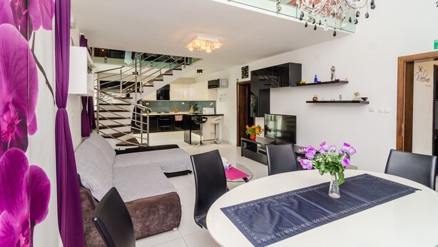 Quarto de hotel moderno com escada de vidro, área de refeições com mesa redonda e sala de estar com sofá e cadeiras.