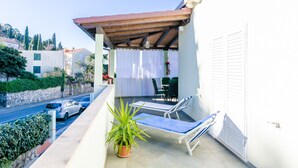 Uma varanda com pérgola, cadeiras de descanso, um vaso de planta e vista para a rua e prédios.