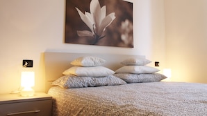Una cama bien hecha con ropa de cama estampada, un cuadro de una flor colgado en la pared y lámparas de noche sobre una mesita.