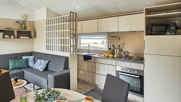 Een moderne woonkamer met een grijze bank, een eettafel met borden en een kopje, en een keuken met een magnetron en fornuis.