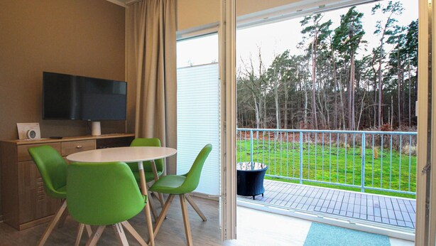 Ein Zimmer mit Fernseher, einem kleinen Tisch und grünen Stühlen. Es besitzt eine Glasschiebetür, die auf einen Balkon mit Blick auf Bäume und eine Rasenfläche führt.