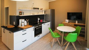 Eine kleine Küche mit Herd, Backofen und Kaffeemaschine, ein Esstisch mit grünen Stühlen sowie ein Fernseher auf einem Holzschrank.