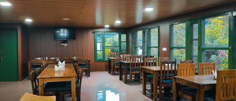 Un restaurante con mesas y sillas de madera, un televisor de pantalla plana en la pared y amplias ventanas con vistas a zonas verdes.