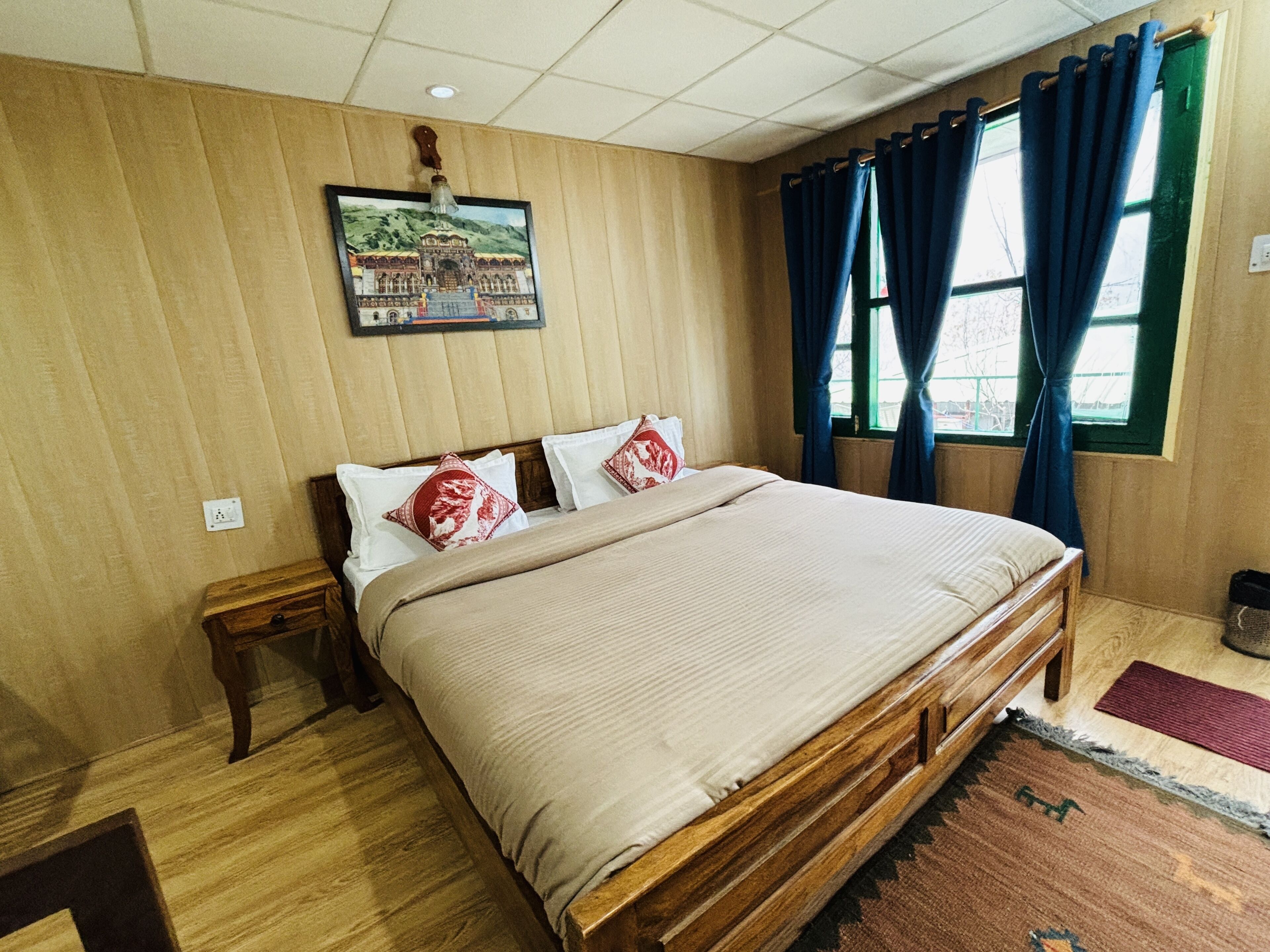 A wooden-paneled room with a large bed, a nightstand, and a window with blue curtains.