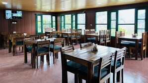 A spacious dining area with wooden tables and chairs, large windows with a view of greenery, and a mounted television on the wall. - Himalayan High, Auli, By Himalayan Eco Lodges (Joshimath)