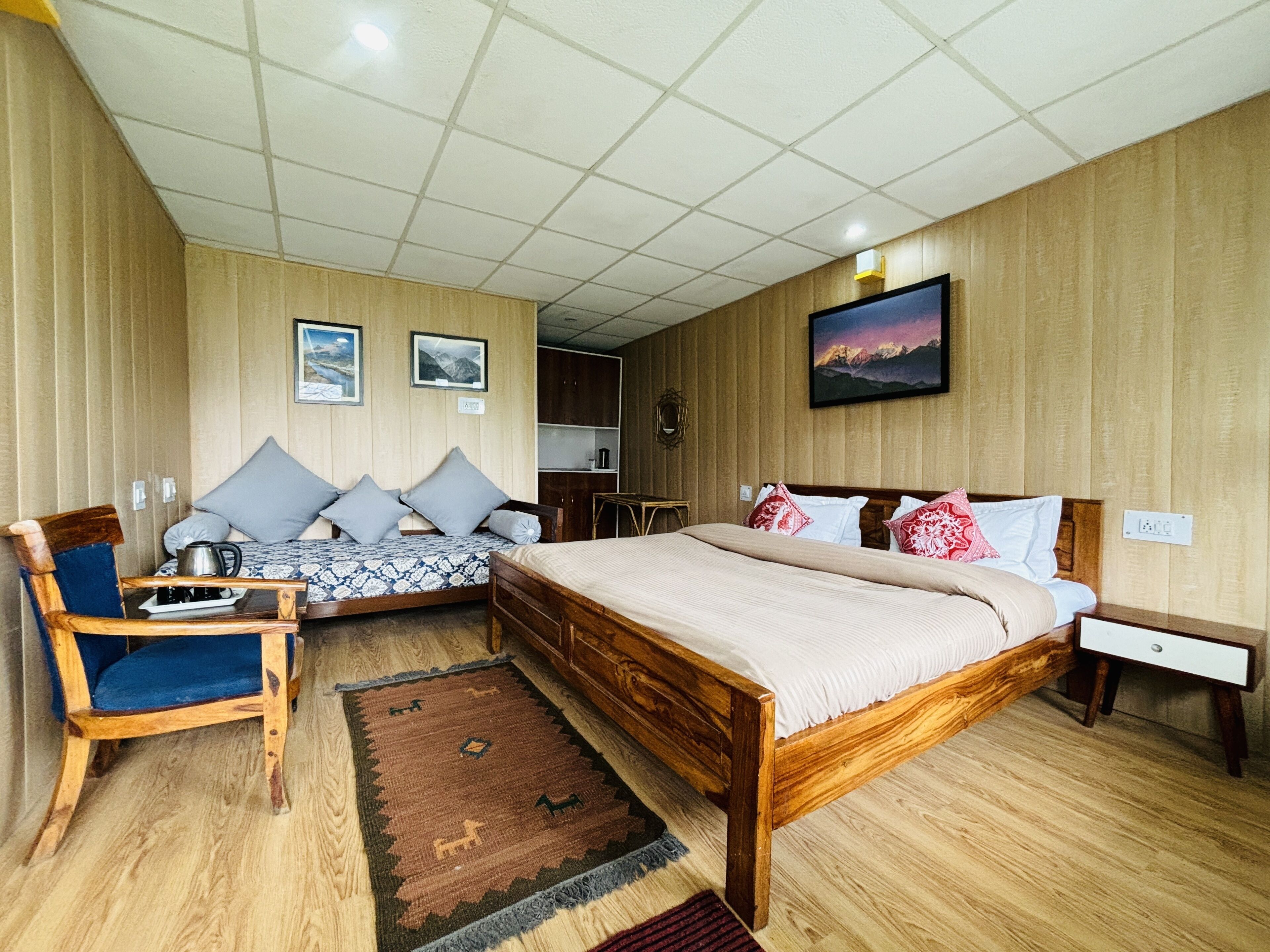 A room with a large bed, a smaller sofa, a wooden chair, and a desk. There are two framed pictures on the wall, a rug on the floor, and a wooden cabinet.