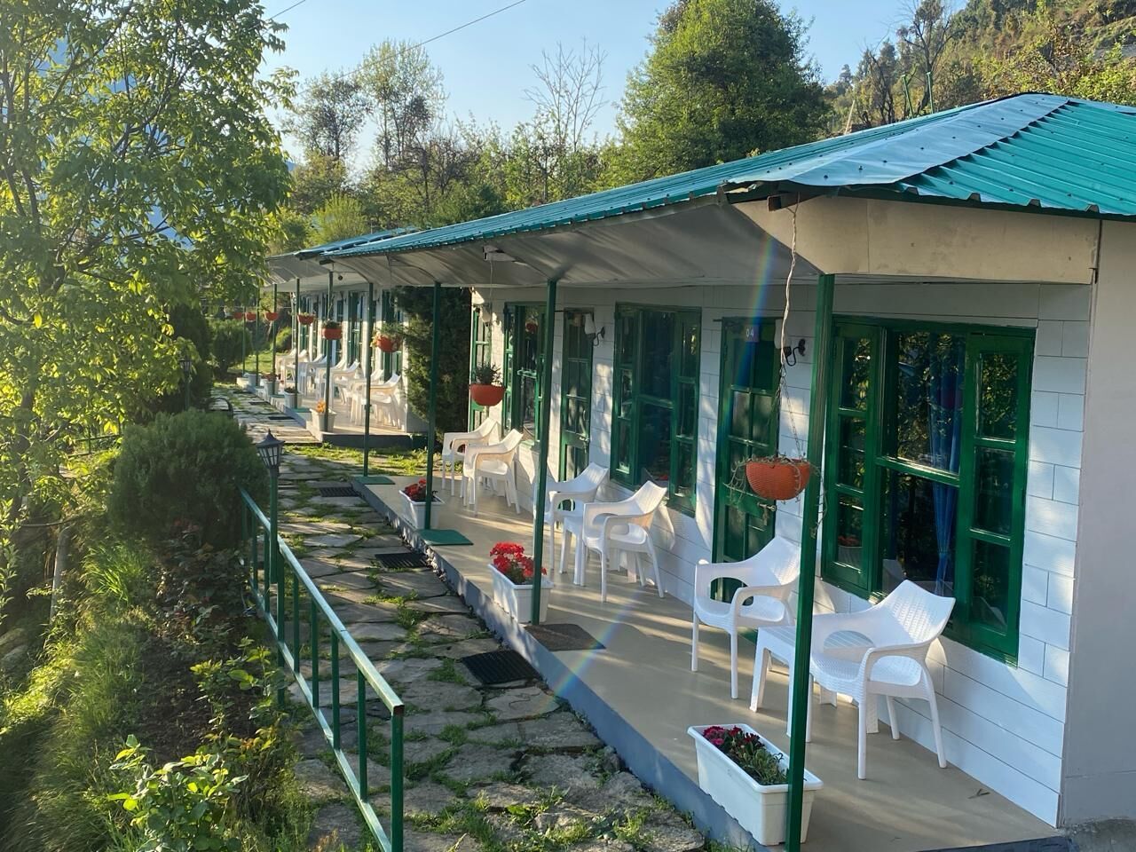 A row of white plastic chairs and tables arranged along a covered outdoor area with green-framed windows.