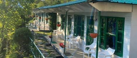 A row of white plastic chairs and tables arranged along a covered outdoor area with green-framed windows.