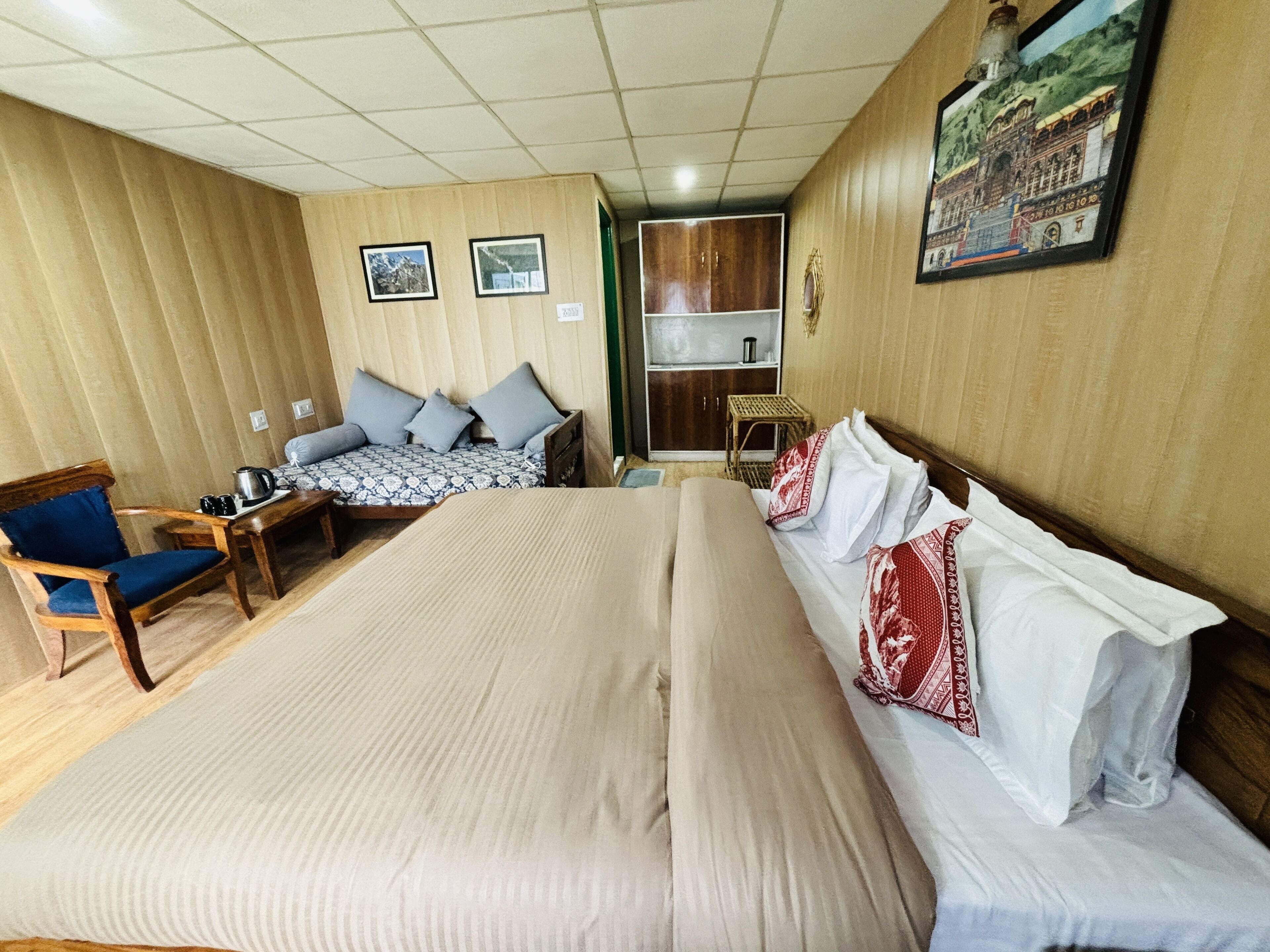 A hotel room with a large bed, a sofa, a wooden chair, a small table with a kettle, and a shelf with a picture frame.