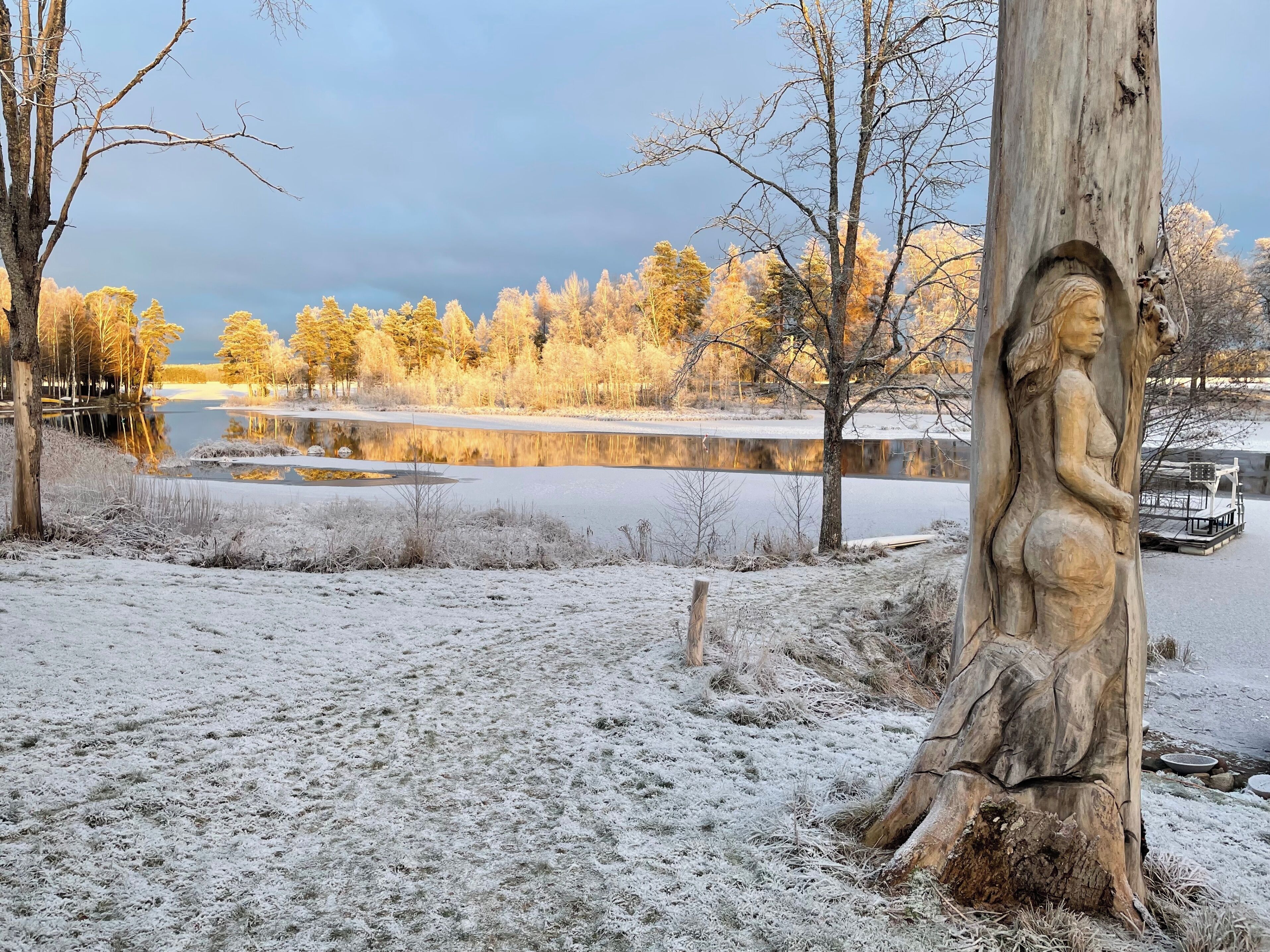 A winter landscape with a frozen lake, trees, and a wooden sculpture.