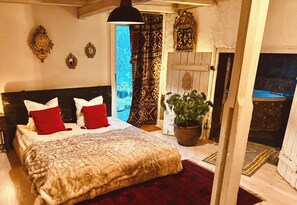 A bedroom with a bed, red pillows, a large potted plant, and a wooden door leading to a stone fireplace. - Romantic Spa Villa With Fireplace by the Lake (Stjärnsund)