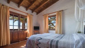 Une chambre avec un lit, des poutres en bois au plafond, une télévision et une vue sur des palmiers.