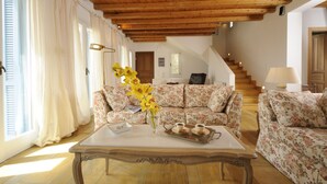 A living room with a wooden ceiling, floral sofas, a coffee table with a vase of flowers, and a staircase in the background.