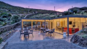 A modern outdoor dining area with glass walls, a concrete floor, and a view of a desert landscape.