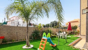 A backyard with a play area, including a slide and a climbing structure, surrounded by a well-maintained lawn and a stone wall. - Apartment "Anastácio" with Garden View, Shared Pool, Wi-Fi & Balcony (Olhos de Água)